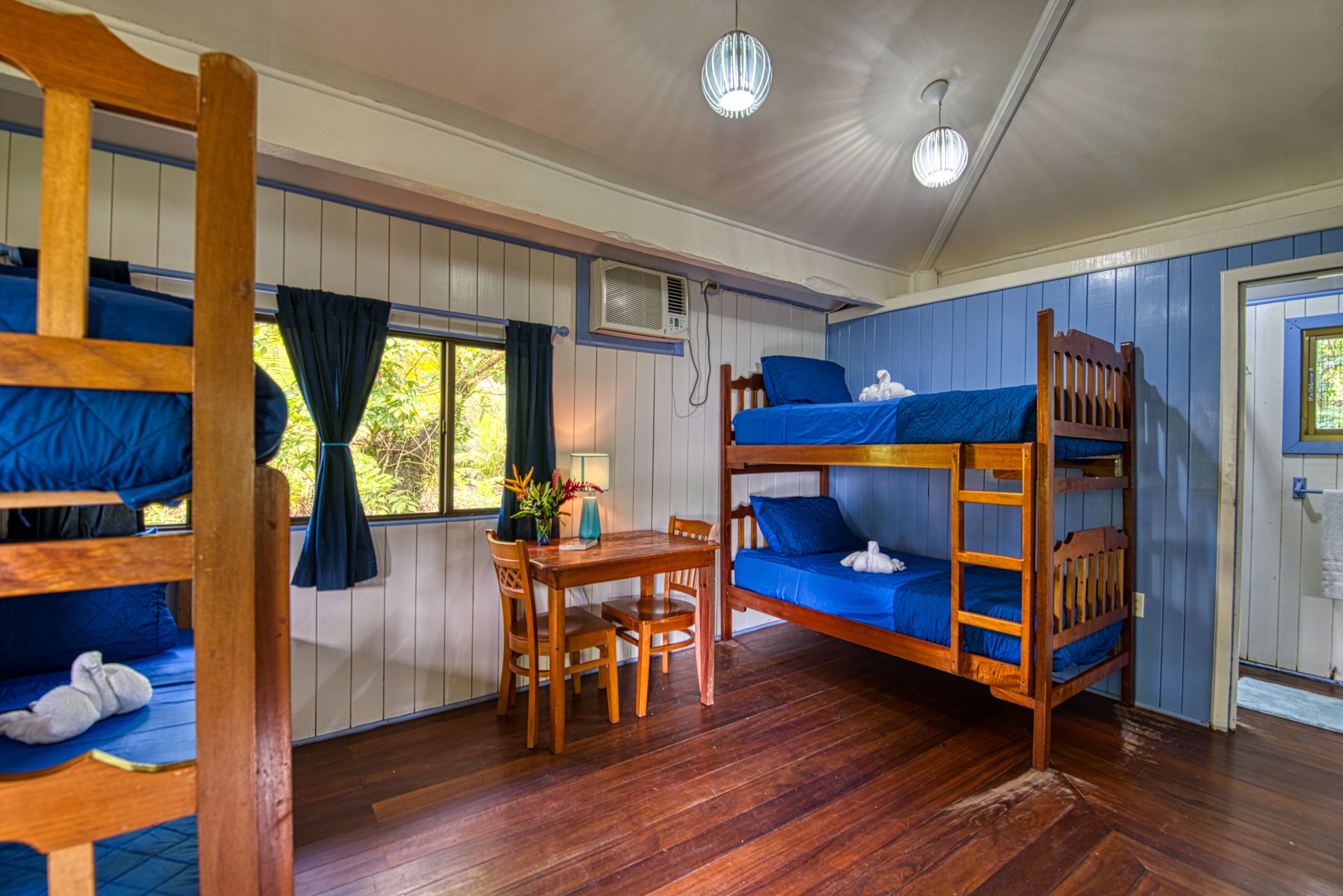 Habitación con literas, una mesa de madera y sillas, una ventana con cortinas y un equipo de aire acondicionado.