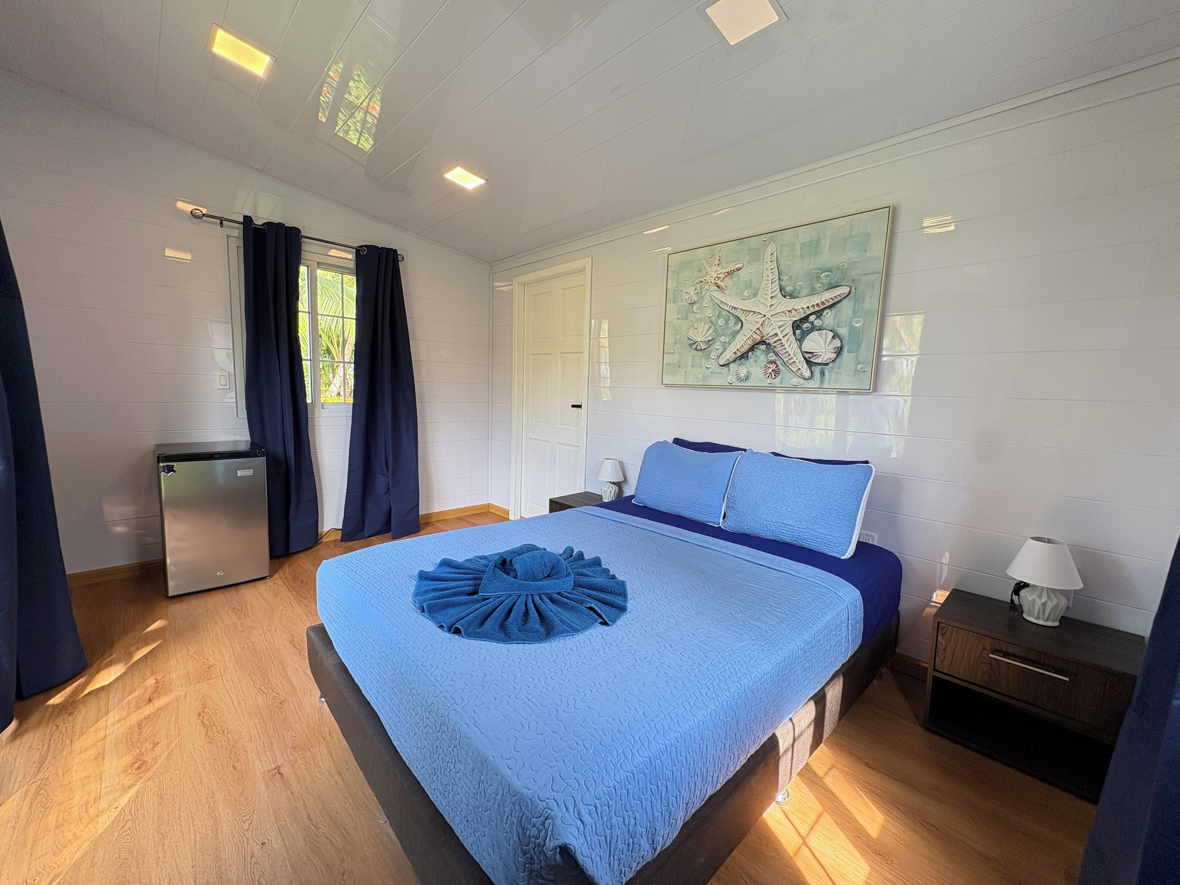 Ein Schlafzimmer mit einem Bett, einem Nachttisch, einer Lampe, einem Fenster mit Vorhängen, einer Tür und einer Wand mit einem Seestern-Kunstwerk.