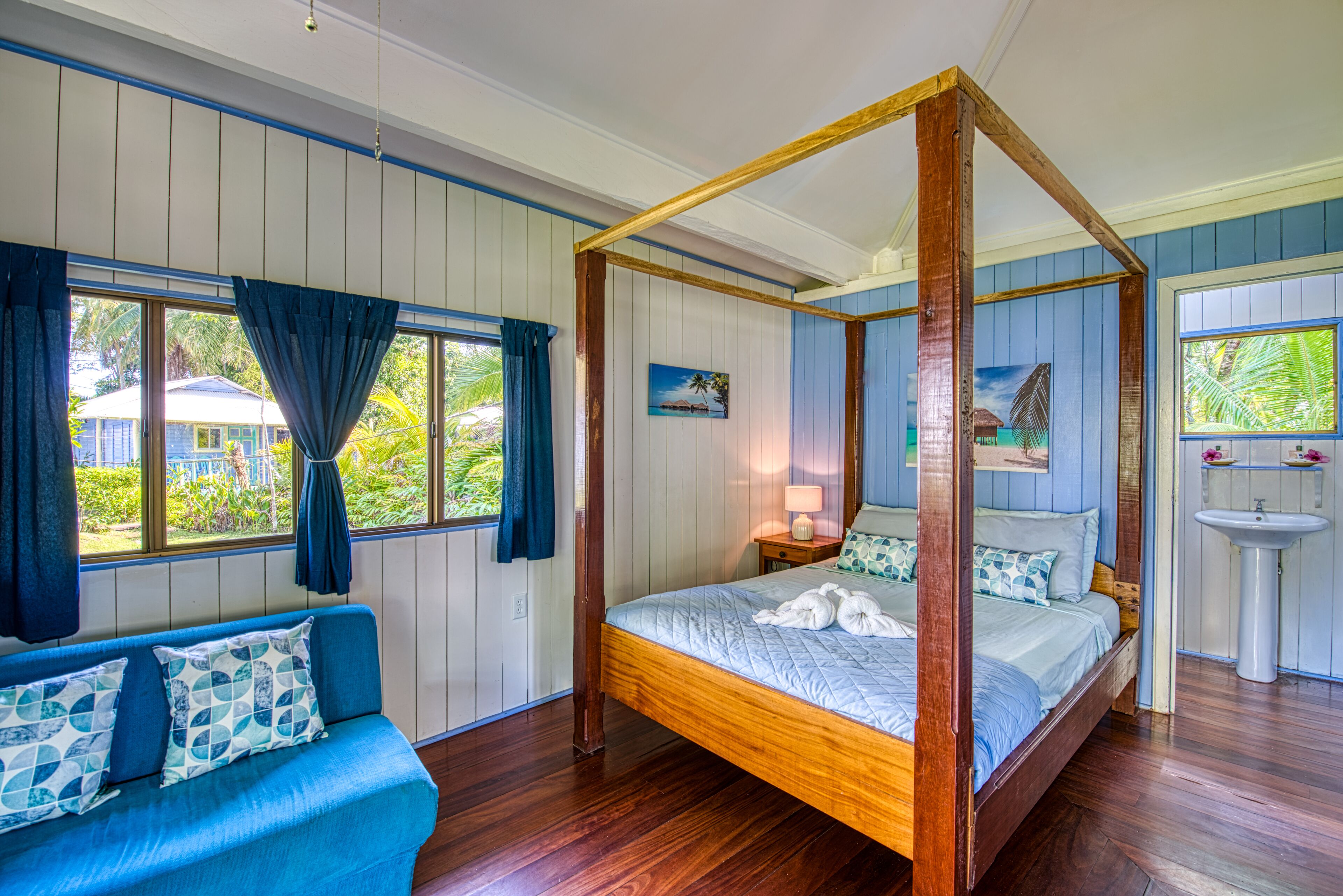 Una cama con dosel de madera, un sofá tapizado en azul con cojines estampados, y vista a una casa a través de una ventana con cortinas azules.