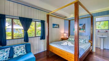 A four-poster bed with a wooden frame, a blue upholstered sofa with patterned cushions, and a view of a house through a window with blue curtains.