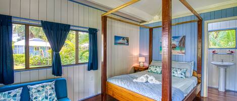 Una cama con dosel de madera, un sofá tapizado en azul con cojines estampados, y vista a una casa a través de una ventana con cortinas azules.