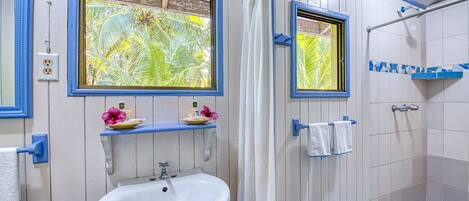 Un baño con un lavamanos blanco, dos ventanas con marcos azules, una ducha con azulejos blancos y azules, y un toallero con dos toallas.