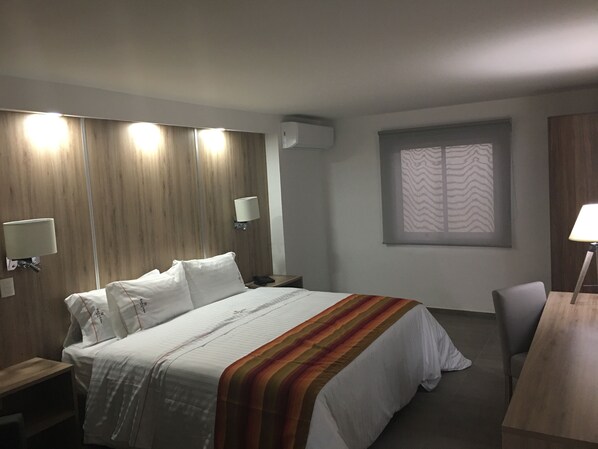 A hotel room with a large bed, bedside lamps, a desk, and a window with a patterned blind. - Hangar Inn Guadalajara Aeropuerto (Tlajomulco de Zúñiga)