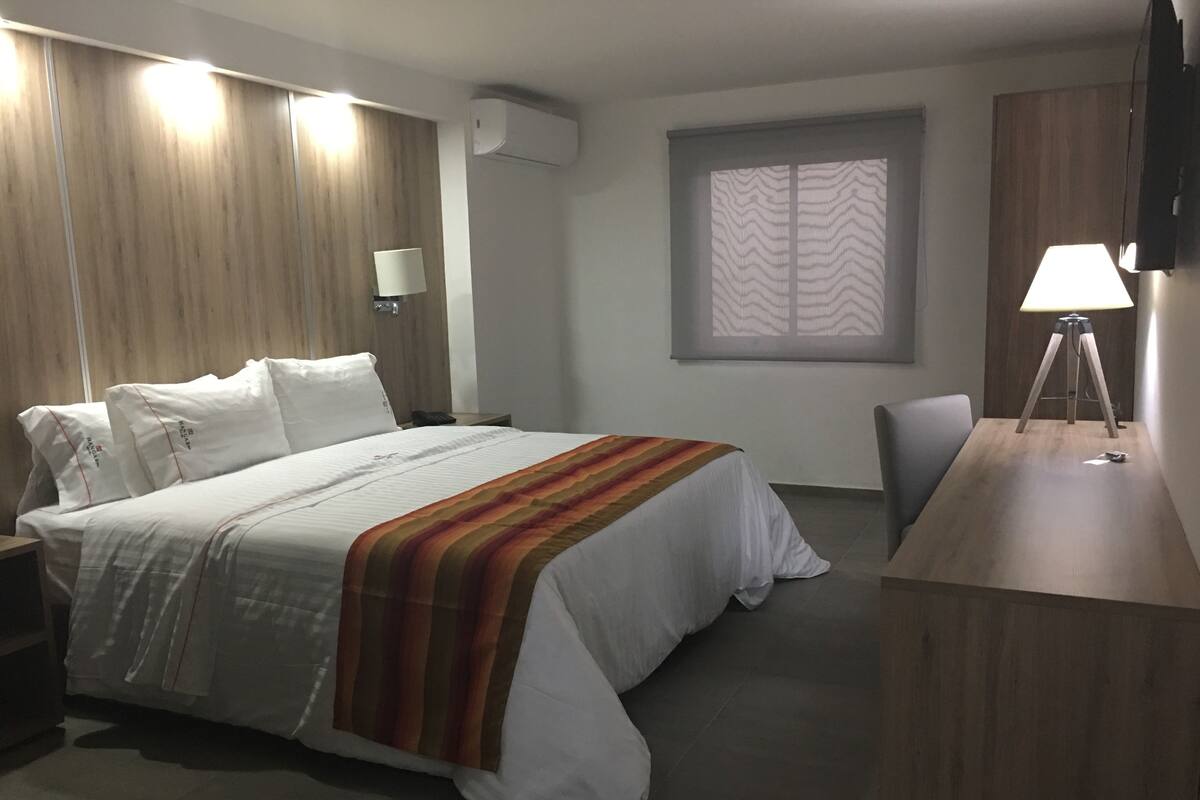 Une chambre d’hôtel comprenant un lit, un bureau, une chaise, une lampe, une fenêtre avec des rideaux à motifs et un appareil de climatisation.