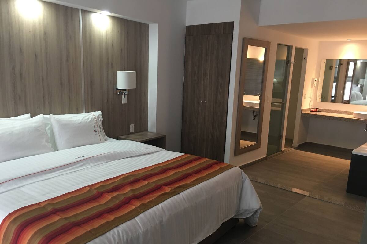 Une chambre d’hôtel avec un grand lit, une table de chevet, un miroir et une salle de bain équipée d’un lavabo et d’un miroir.