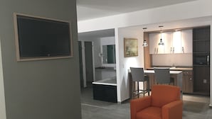 A modern living room with a large flat-screen TV, a dining area with a kitchen in the background, and an orange armchair.