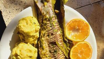 Ein ganzer Fisch in Blätter gewickelt, serviert mit Kartoffelpüree und einer Orangenscheibe auf einem Teller.