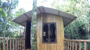 Una cabaña de madera con techo de metal, rodeada por una terraza y barandillas de madera, ubicada en zona boscosa.