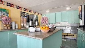 A kitchen with teal cabinets, a central island with a bowl of fruit, a coffee maker, and a microwave. - Kings Inn Anaheim (Anaheim)
