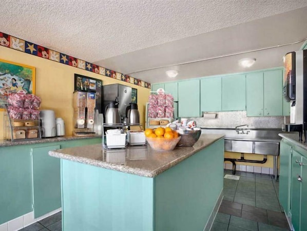 A kitchen with teal cabinets, a central island with a bowl of fruit, a coffee maker, and a microwave. - Kings Inn Anaheim (Anaheim)