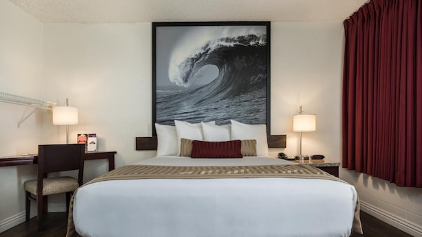 A hotel room with a large bed, two bedside tables with lamps, a desk with a chair, and a large wave painting on the wall. - Kings Inn Anaheim (Anaheim)