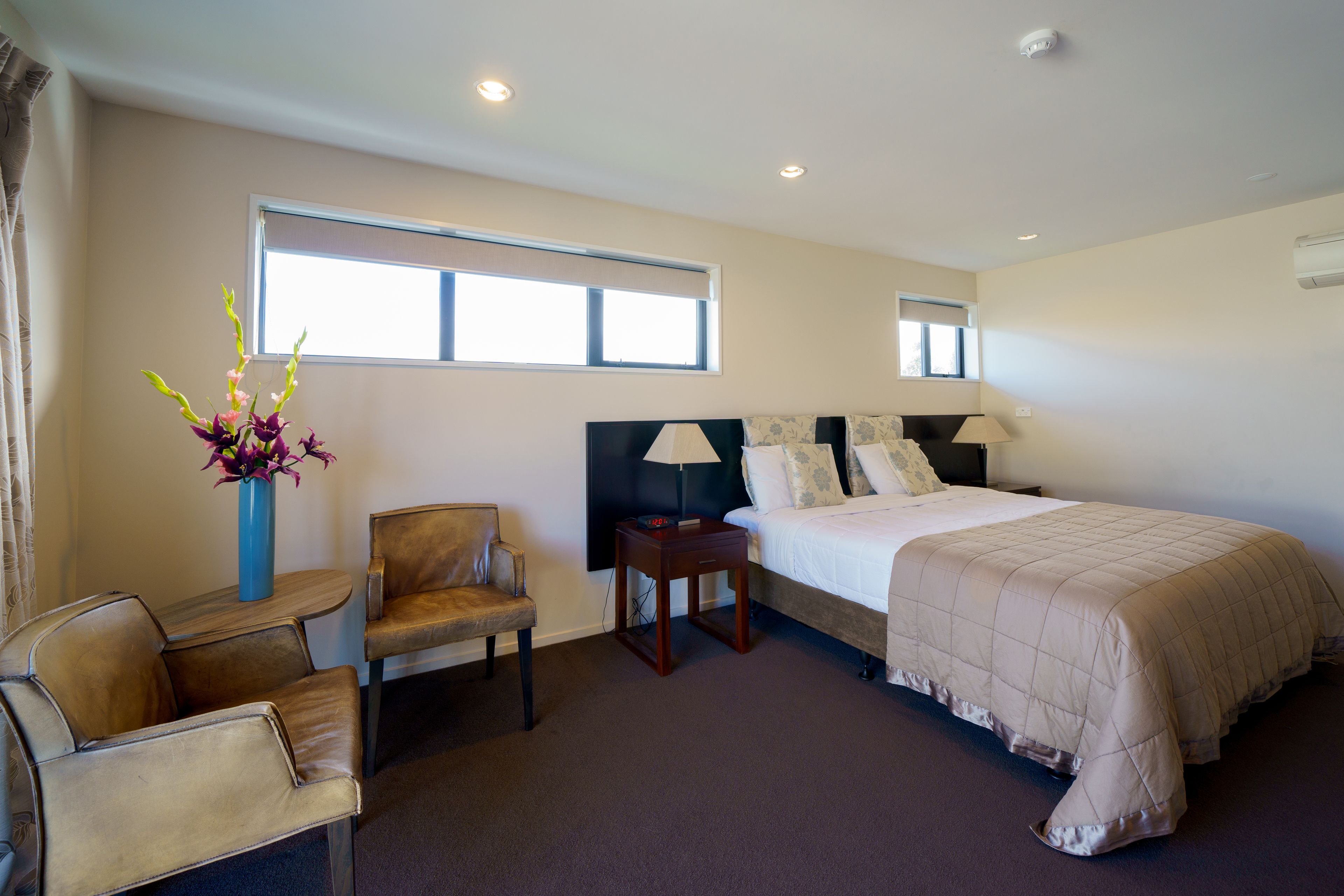 A hotel room with a bed, a chair, a small table, a vase with flowers, and a window.