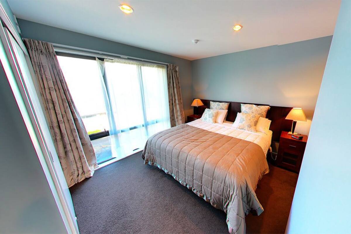 A hotel room with a large bed, two bedside tables with lamps, a view through a sliding door to a balcony, and a carpeted floor.
