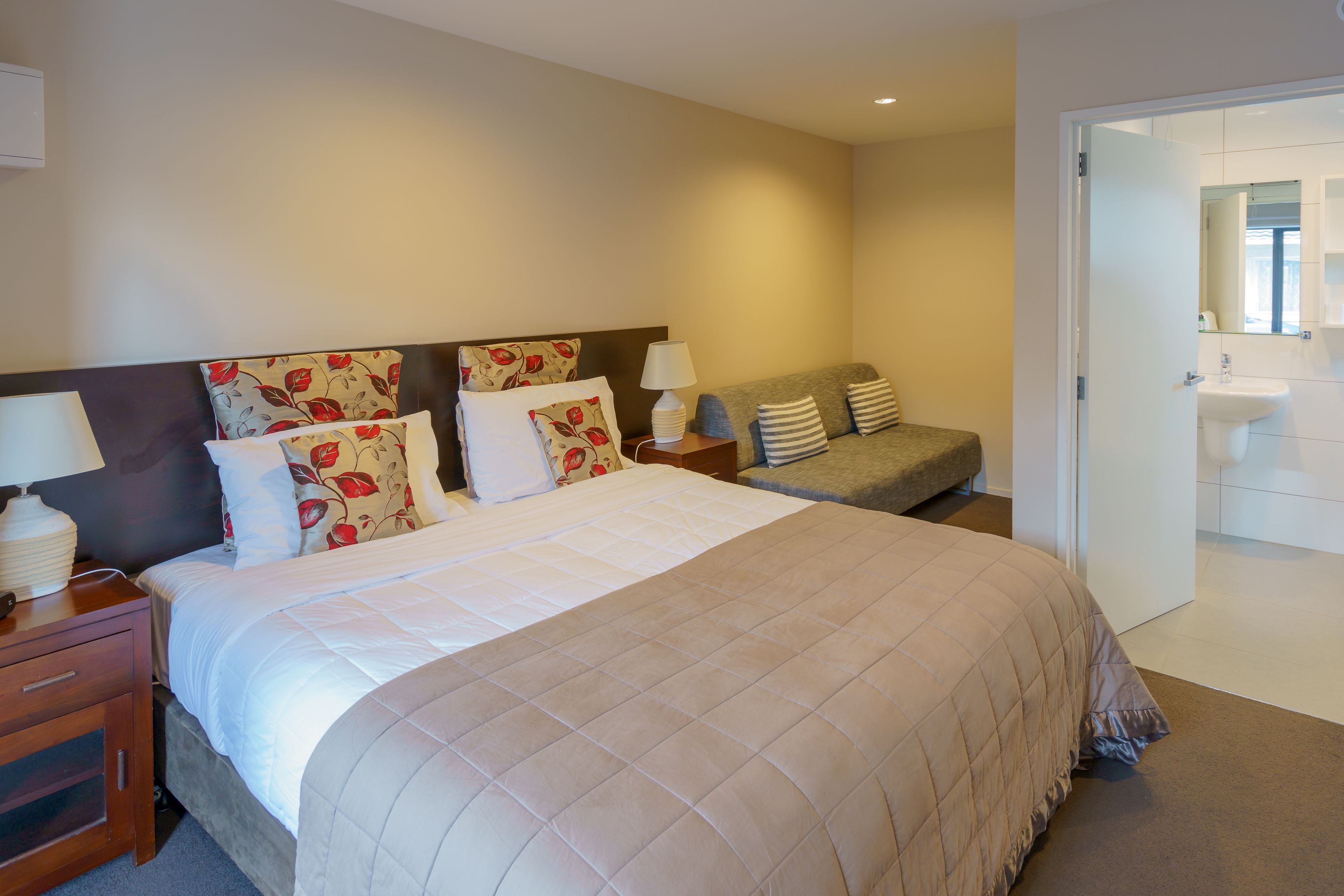 A hotel room with a large bed, a bedside table with a lamp, a sofa, and a bathroom visible through an open door.