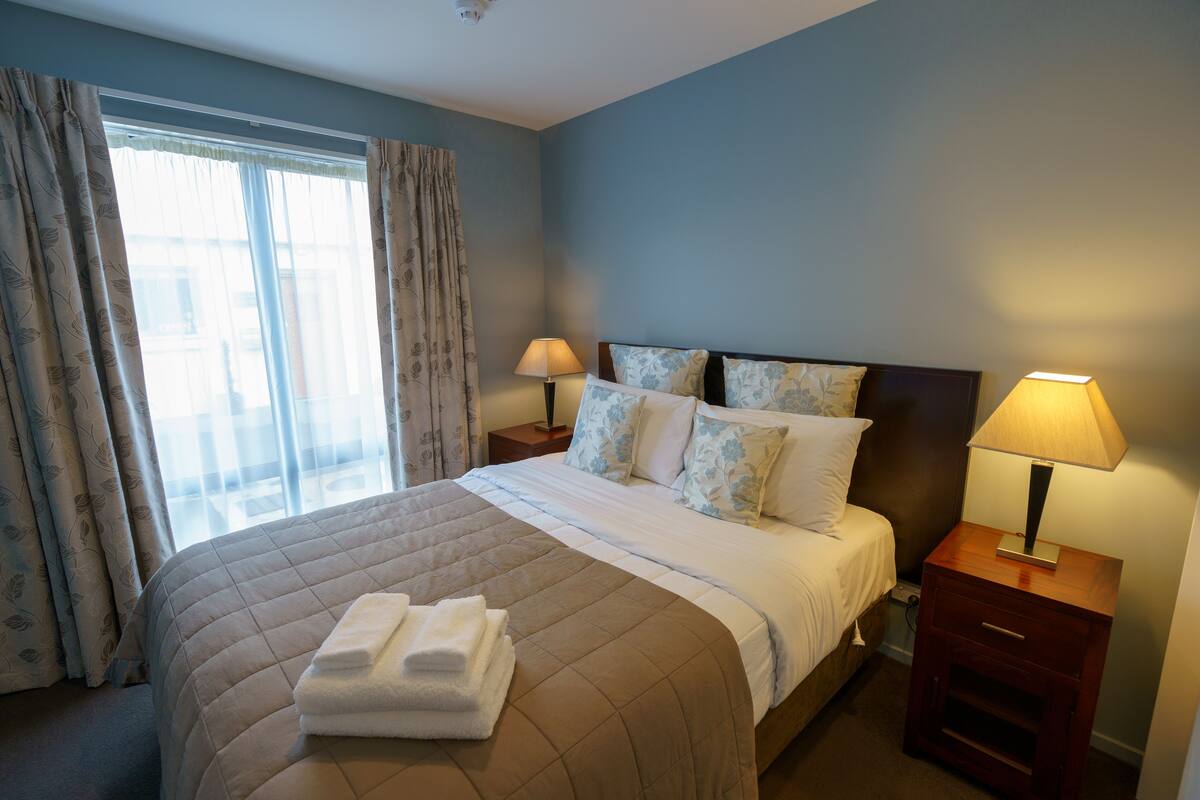 A neatly made bed with a brown quilt, two bedside lamps, and a window with curtains.