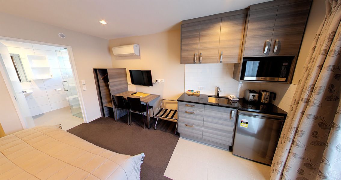 Une chambre d’hôtel compacte comprenant un lit, un bureau avec une chaise, une kitchenette et une salle de bain avec douche.