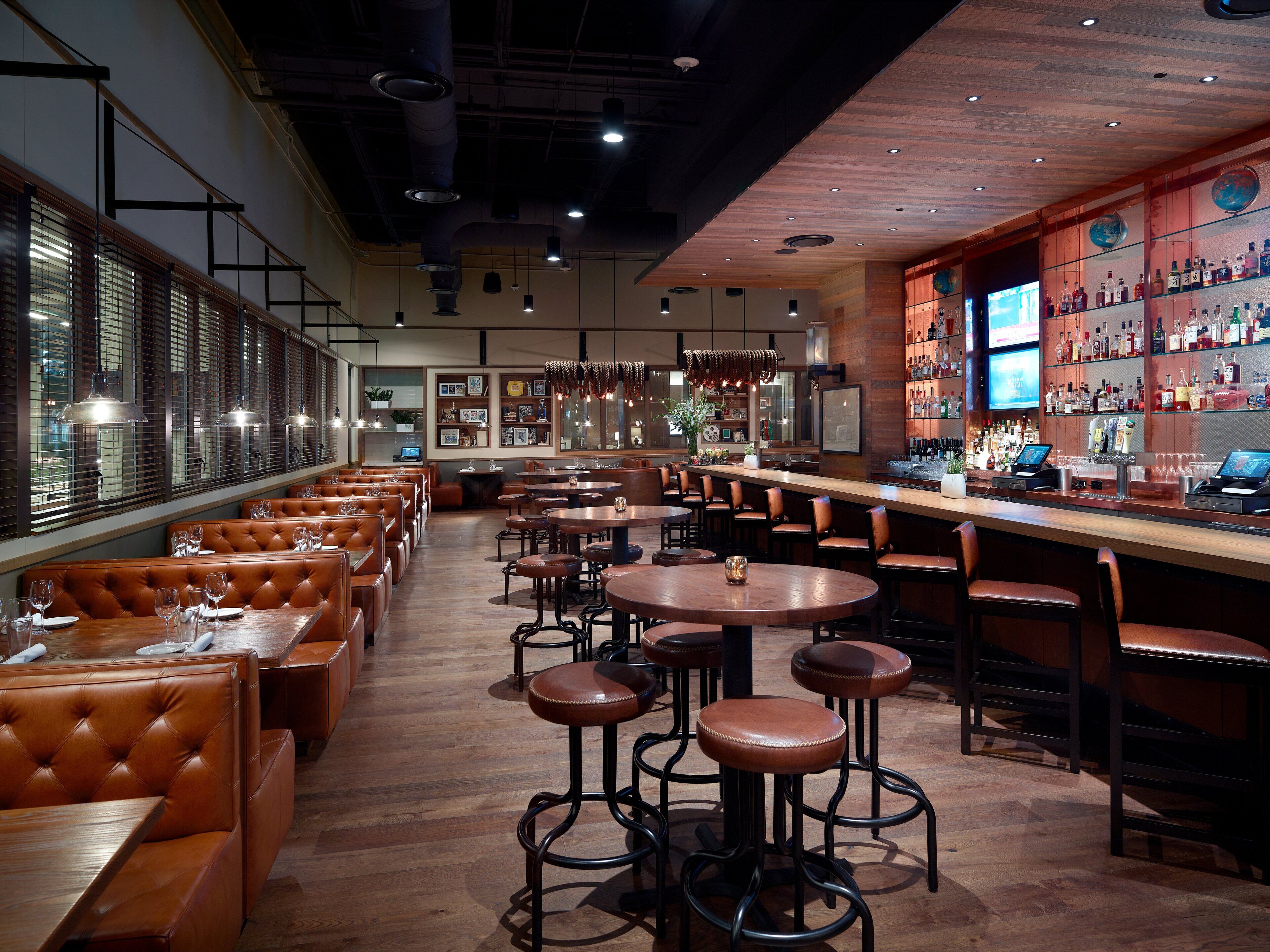 A restaurant with booths, high tables, and bar stools. There are multiple tables and chairs, and a bar area with a variety of bottles.