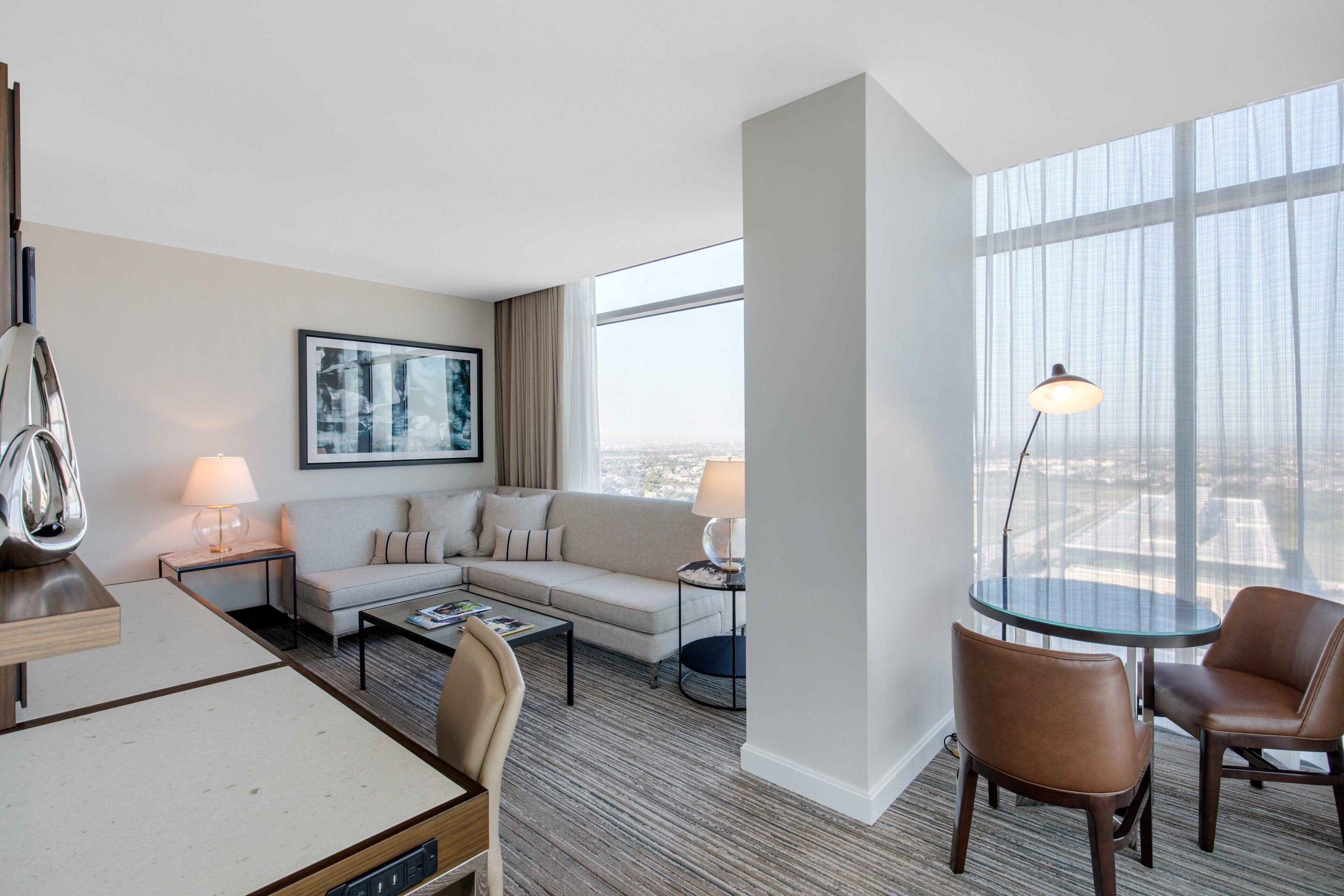 A modern hotel room with a sofa, coffee table, and chairs, a glass table, and a large window offering a city view.