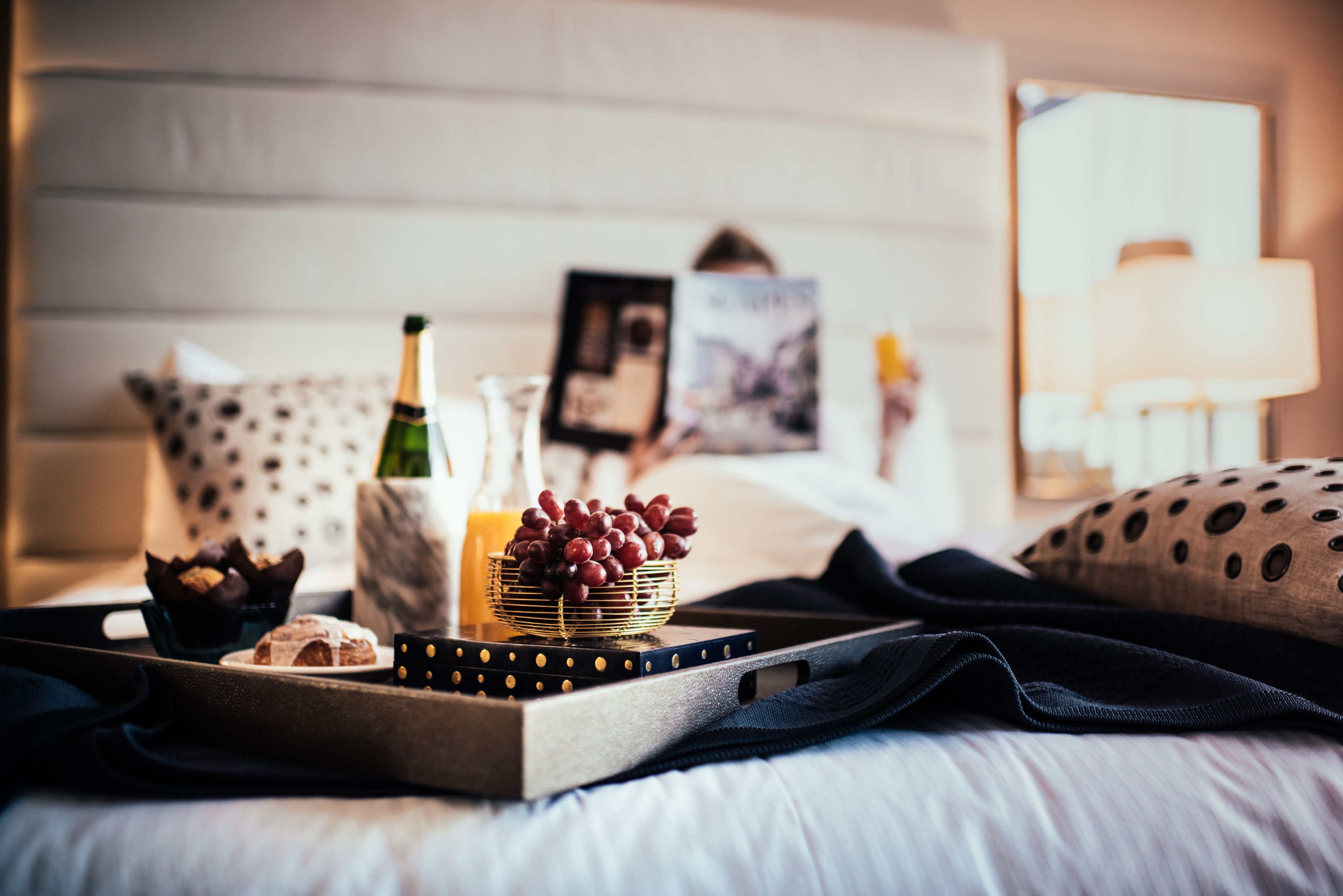 A neatly arranged breakfast tray on a bed with a polka dot pillow, a bottle of champagne, a bowl of grapes, and a muffin.