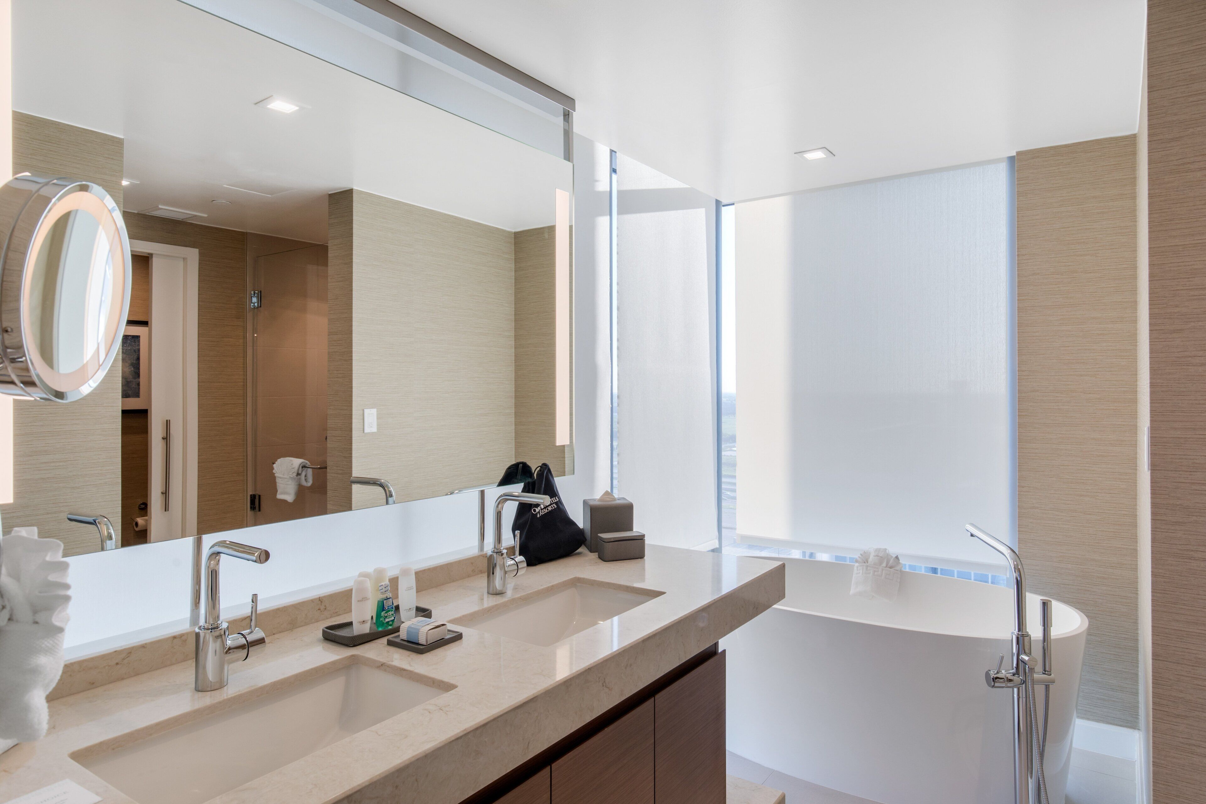 A modern bathroom with a large mirror, two sinks, a bathtub, and a window providing natural light.
