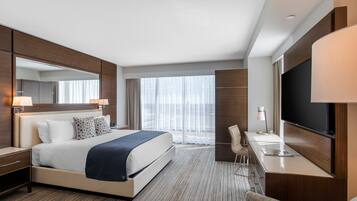 Una habitación de hotel moderna con una cama grande, un escritorio, un televisor de pantalla plana, una silla y un amplio ventanal con cortinas.