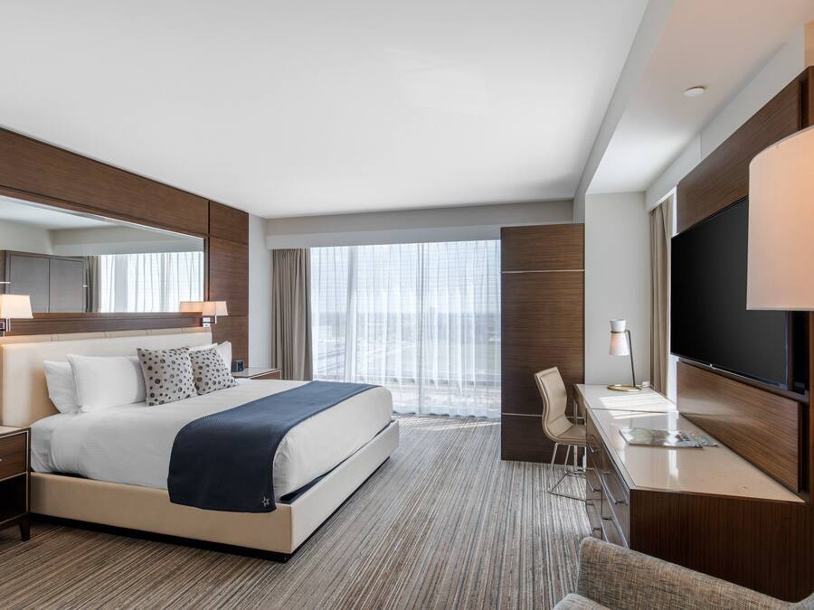 Una habitación de hotel moderna con una cama grande, un escritorio, un televisor de pantalla plana, una silla y un amplio ventanal con cortinas.