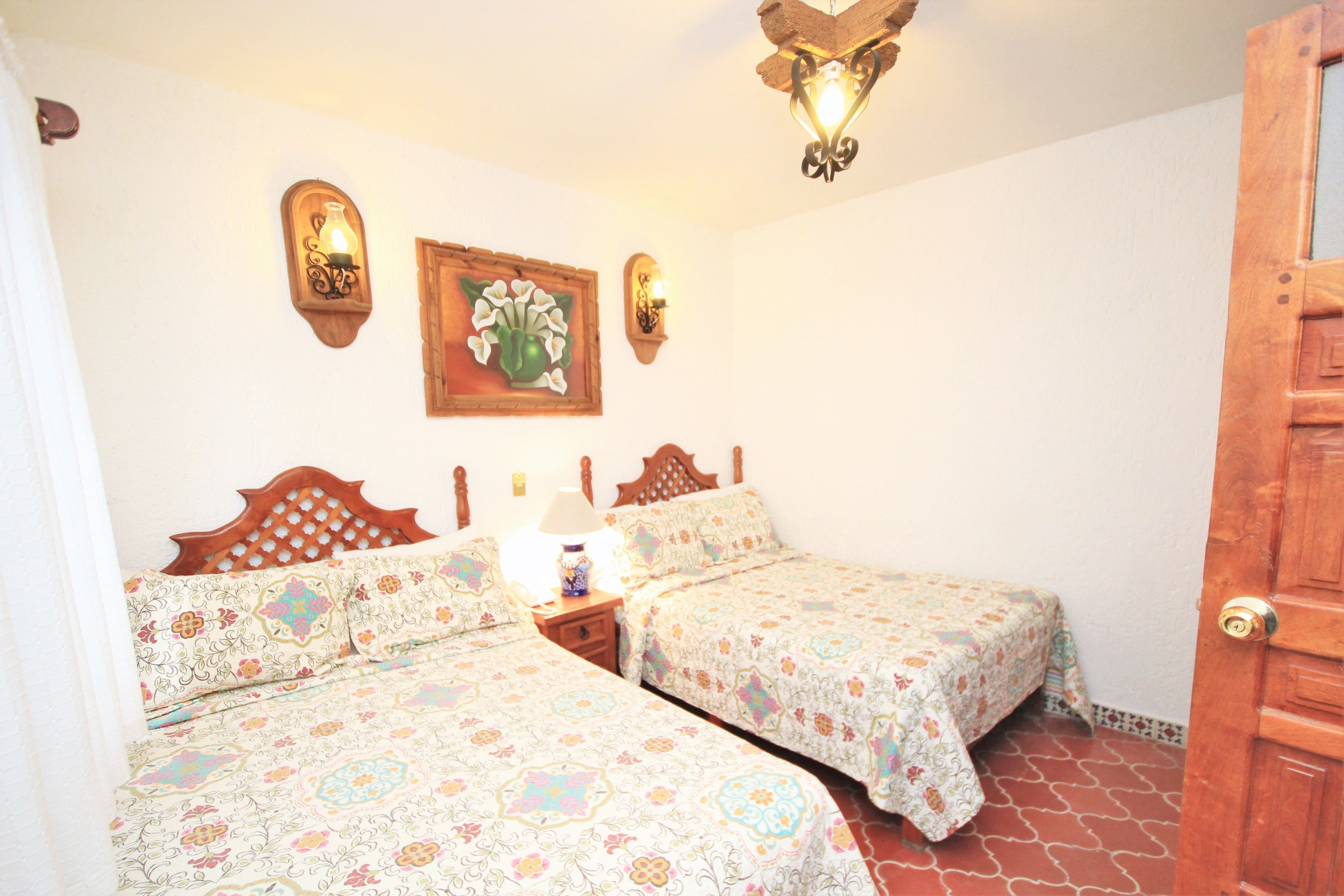 Una habitación con dos camas, cabeceros de madera, una mesita de noche con lámpara y una puerta de madera.