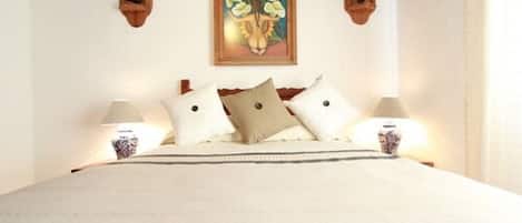 A neatly made bed with white linens and two pillows, a framed artwork above the bed, and two wall-mounted lamps on either side.