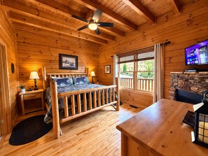Un dormitorio de cabaña de madera con cama, mesitas de noche, un escritorio, chimenea y televisor.
