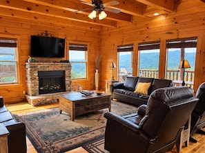 A log cabin interior with a stone fireplace, leather furniture, and a television mounted on the wall.
