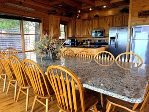 A wooden cabin interior with a granite dining table surrounded by wooden chairs, a kitchen area with stainless steel appliances, and a welcome sign.
