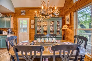 Interior de una cabaña de madera con una mesa de comedor preparada para cuatro personas, rodeada de sillas, una araña y un aparador con puertas de cristal que exhibe vajilla.