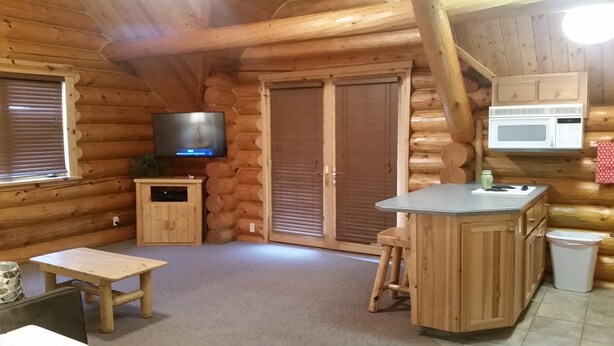 Interior de una cabaña de troncos con televisor, armarios de madera, una zona de cocina y una mesa de comedor.