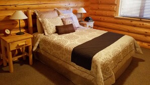 Un dormitorio de cabaña de troncos con una cama, dos mesitas de noche, lámparas y una ventana con persianas.