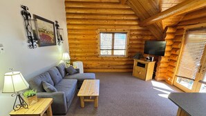 Interior de una cabaña de troncos con un sofá, una mesa de madera, un televisor y una ventana con persianas.