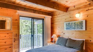 A wooden cabin bedroom with a bed, a nightstand, a lamp, a framed picture, and a sliding glass door leading to a balcony.