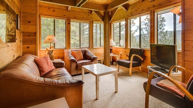 A wooden cabin interior with a sofa, chairs, a table, a TV, and large windows.