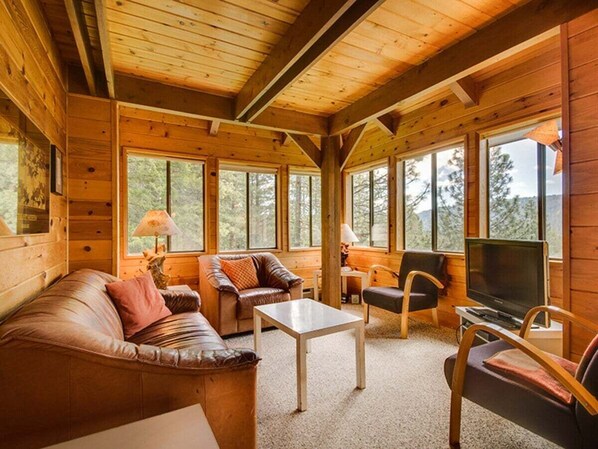 Interior de una cabaña de madera con sofá, sillas, una mesa, televisor y ventanas amplias.