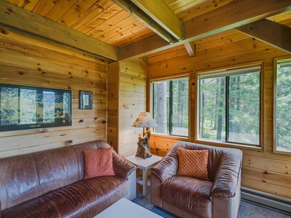 Interior de una cabaña de madera con un sofá de cuero marrón, una mesita con una lámpara y ventanas grandes.