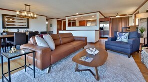A modern living room with a brown leather sofa, a blue armchair, and a coffee table. There is a dining area with a wooden table and chairs, and a kitchen with a refrigerator and cabinets.