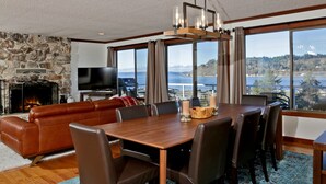 Una sala de estar moderna con chimenea de piedra, un televisor de pantalla plana grande, una mesa de comedor de madera y vistas a un lago y montañas.