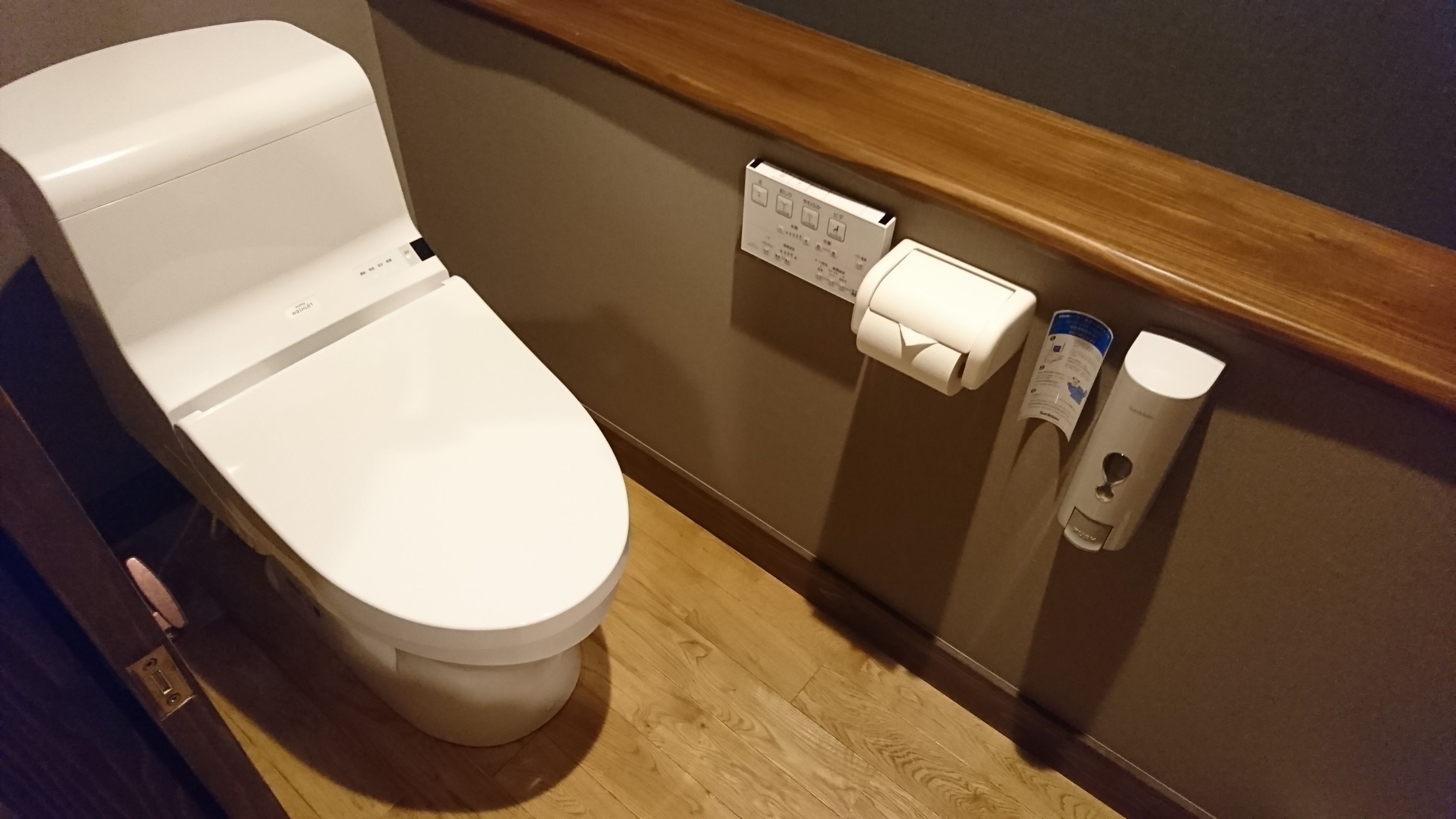 A white toilet with a flush handle, a paper towel dispenser, and a soap dispenser on the wall.