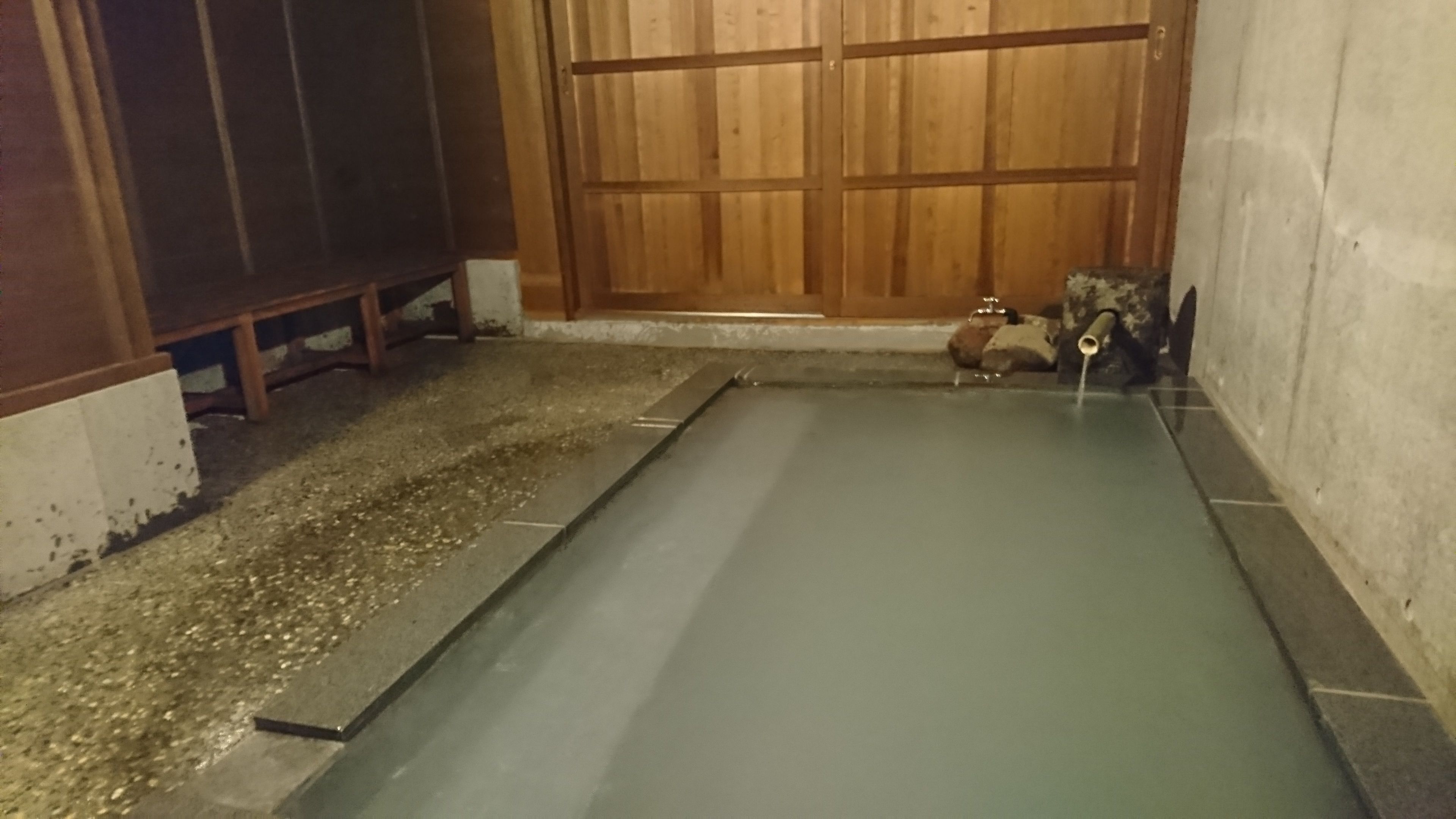 A small, enclosed hot spring area with a wooden door, a stone bench, and a small water feature.