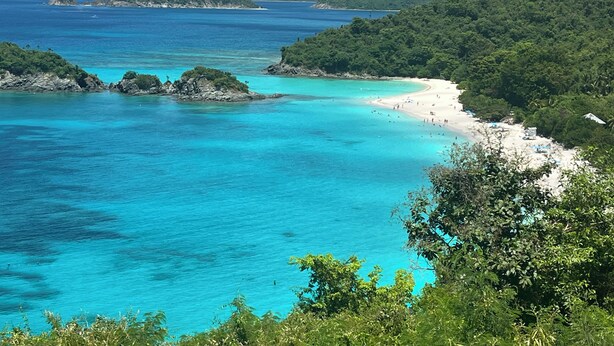 Paesaggio costiero con spiaggia, acque turchesi cristalline e colline verdeggianti sullo sfondo.