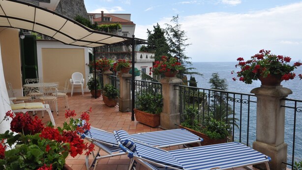 Eine Terrasse mit zwei Liegestühlen, einem Tisch mit Stühlen, Topfpflanzen und Blick auf das Meer.