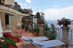 A terrace with two sun loungers, a table with chairs, potted plants, and a view of the sea.