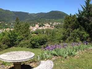 Un tavolo e una panchina in pietra in un giardino con fiori viola e vista su un villaggio in lontananza.