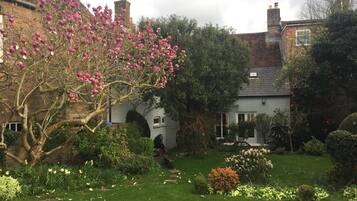 Un giardino con diverse piante e fiori, un edificio in mattoni con un camino e un cielo sereno.
