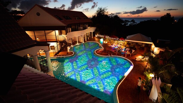 A swimming pool with a unique design, a staircase leading to a covered area, and a building with a red roof. - The Pool Resort Villa Hasta Manana (Onna)