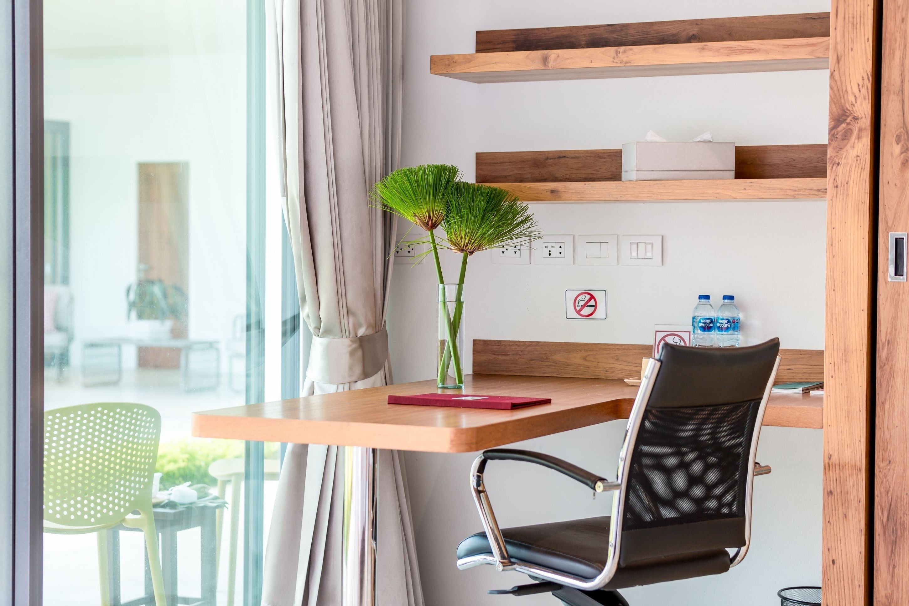 A modern room with a wooden desk, a chair, a vase with green plants, and a window with curtains.